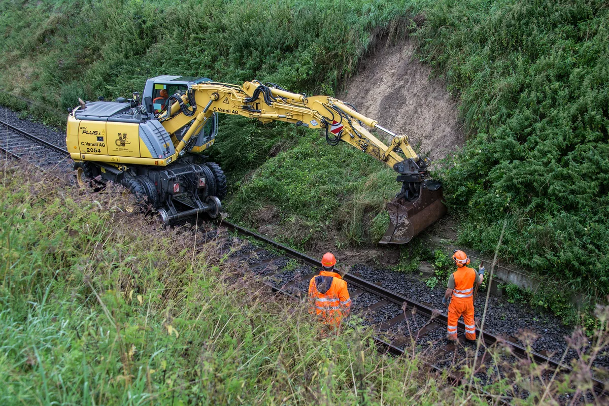 Ein Schienenbagger räumt einen Erdrutsch von den Schienen im Weiler Fistel weg. Er wird dabei von zwei Arbeitern beobachtet.
